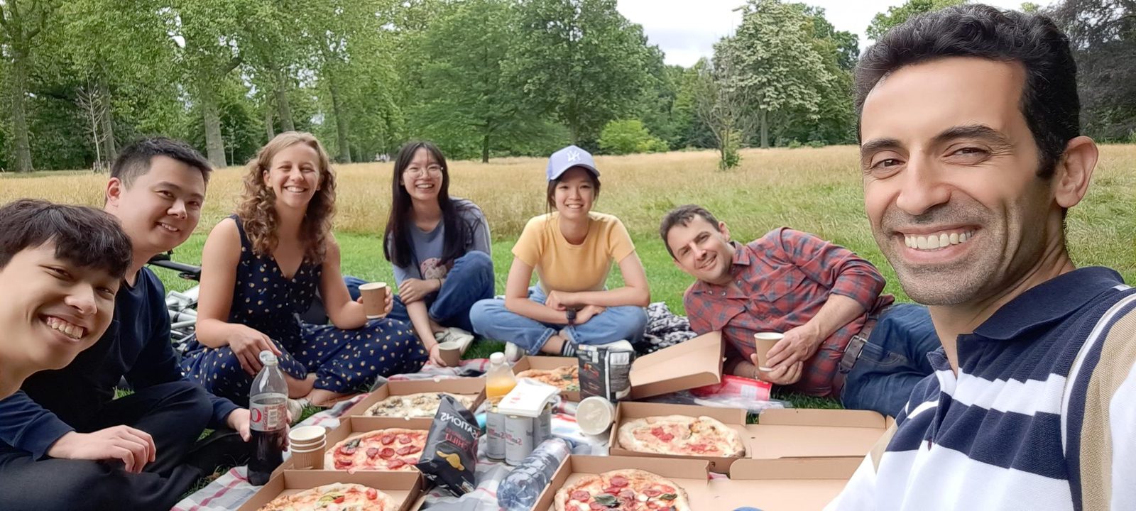 The lab sitting on the grass in a park around pizza boxes and drinks.