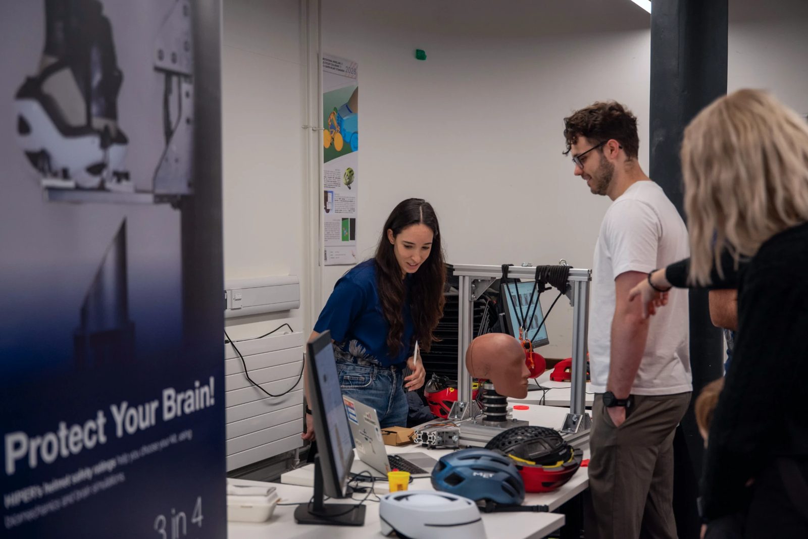 HEAD Lab members talking to visitors at the Great Exhibition Road Festival, beside a “Protect Your Brain!” banner and a helmet impact rig.